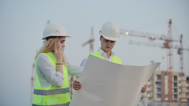 Woman-talking-on-the-phone-and-asks-the-Builder-what-is-on-the-drawings-standing-on-the-background-of-buildings-under-construction
