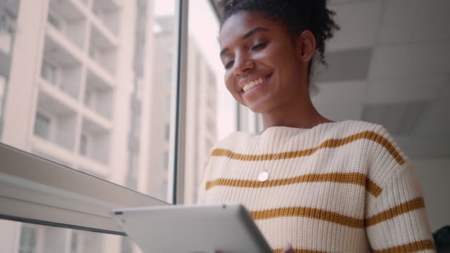 Porträt-einer-glücklichen-jungen-afrikanischen-Geschäftsfrau,-die-mit-einem-digitalen-Tablet-in-der-Nähe-des-Bürofensters-steht
