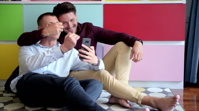 Gay-couple-using-phone-on-arm-chair-together.-Showing-ring.