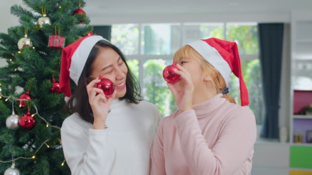 Asian-Lesbian-couple-celebrate-Christmas-festival.-LGBTQ-female-teen-wear-Christmas-hat-relax-happy-play-Christmas-ball-enjoy-xmas-winter-holidays-together-in-living-room-at-home.-Slow-motion-shot.