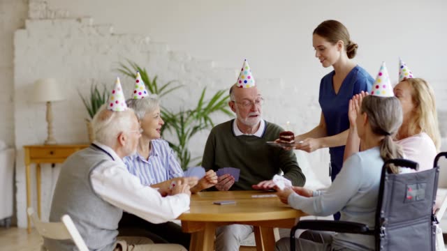 Nurse-bringing-piece-of-birthday-cake-for-senior-man-playing-cards-with-aged-friends-in-party-hats-in-nursing-home.-Aged-man-receiving-wishes-and-kisses-and-blowing-candle-out,-tracking-shot