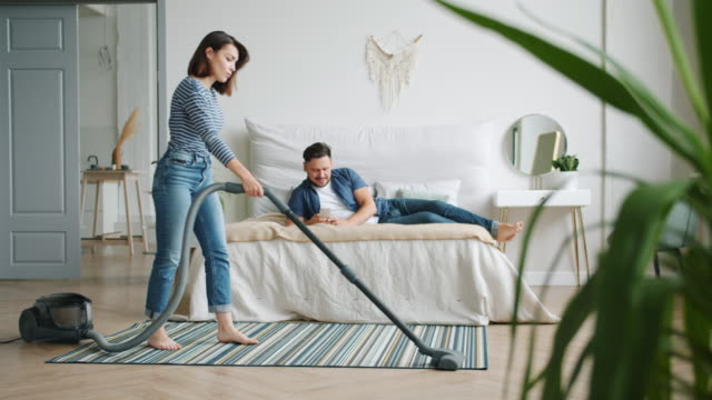 Frau-staubt-Teppich-im-Schlafzimmer,-während-Mann-mit-Smartphone-im-Bett