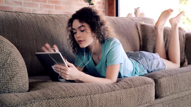 Hispanische-lockige-Haare-junge-Frau-surfen-im-Netz-mit-digitalen-Tablet-und-Musik-hören,-während-sie-auf-der-Couch-entspannen