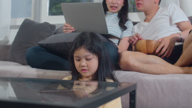 Joven-familia-asiática-e-hija-feliz-usando-la-tableta-y-la-computadora-portátil-en-casa.-Madre-japonesa,-padre-relajarse-con-niña-viendo-películas-y-comprobar-las-redes-sociales-tumbado-en-el-sofá-en-la-sala-de-estar.