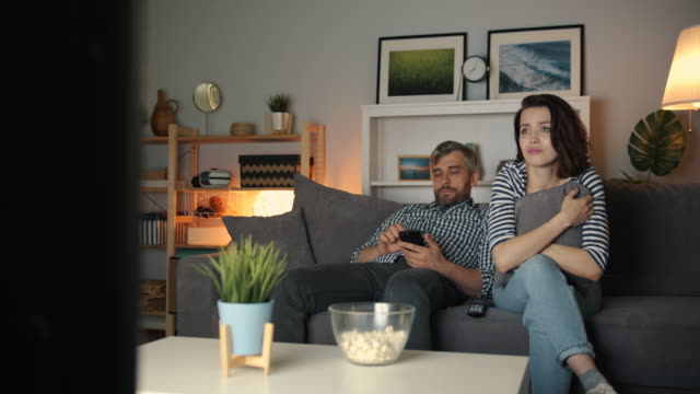 Woman-watching-TV-crying-while-guy-using-smartphone-at-home-at-night
