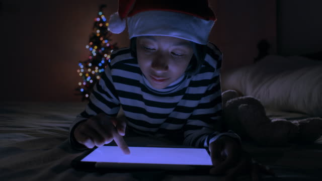 Mittlerer-Schuss.-Asiatische-von-kleinen-Mädchen-mit-digitalen-Tablet,-während-auf-dem-Bett-im-Schlafzimmer-mit-Weihnachtsbaum-in-der-Nacht-dekoriert-liegen