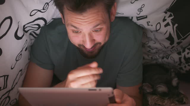 Close-up-of-face-of-a-young-bearded-and-mustashed-man,-using-digital-tablet-under-blanket,-in-his-bed,-at-night,-and-rubs-his-face-and-eyes-out-of-fatigue.-His-yorkshire-terrier-is-lying-next-to-him