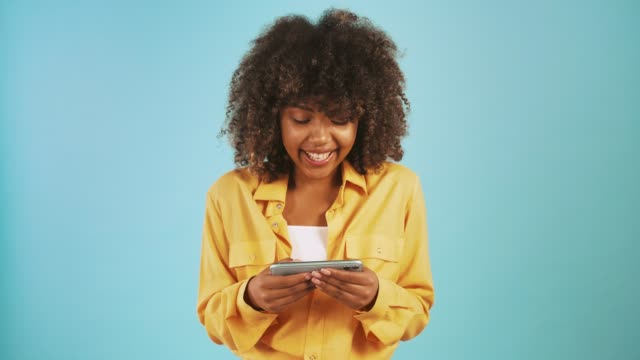 Afro-amerikanische-weibliche-Spielen-Videospiel-auf-dem-Smartphone-und-lächelnd,-während-posiert-vor-blauem-Studio-Hintergrund