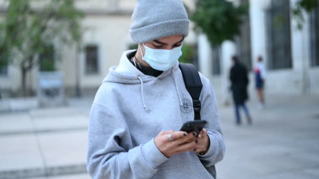 Student-in-a-mask-on-the-street-with-a-smartphone.-Portrait-of-young-man-in-casual,-covid-19,-backpack-and-medical-mask-standing-and-using-smartphone,-chatting-with-friends,-texting,-scrolling
