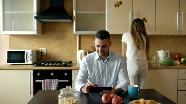 Glücklich-Jüngling-mit-digitalen-Tablettcomputer-sitzen-in-der-Küche,-während-seine-Freundin-zu-Hause-kochen