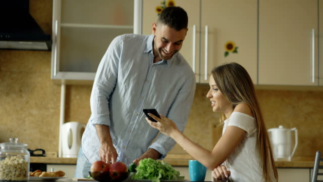 Attraktives-Paar-in-der-Küche-am-frühen-Morgen.-Schöne-Mädchen,-die-gemeinsame-Nutzung-von-social-Media-auf-Smartphone-mit-ihrem-Freund