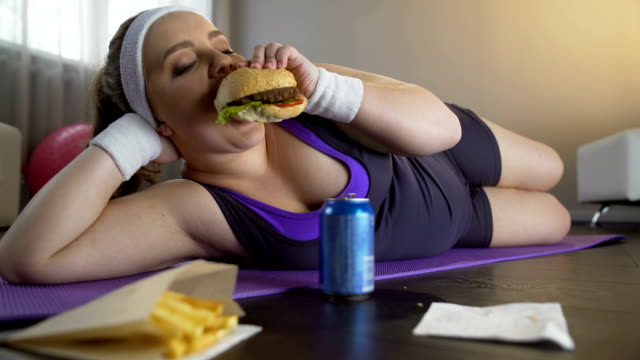 Happy-self-confident-obese-girl-eating-greasy-burger-instead-of-sports-workout