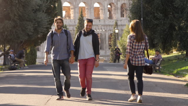 Young-happy-gay-couple-tourists-walk-in-park-road-with-trees-colosseum-in-background-in-rome-at-sunset-holding-hands-lovely-slow-motion-colle-oppio