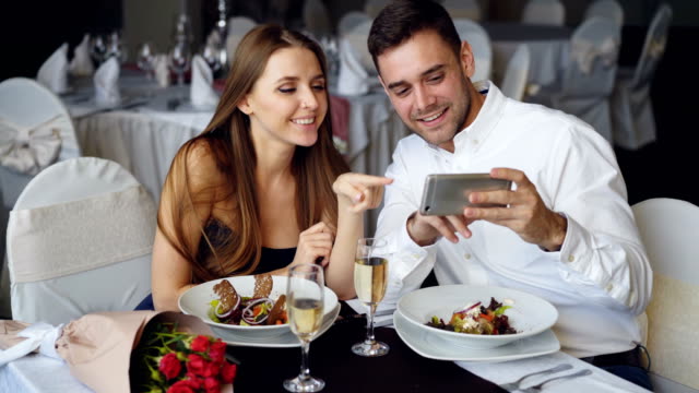 Glückliche-attraktive-junge-Menschen-beobachten-Smartphone-zusammen,-lächelnd-und-beim-Abendessen-im-Restaurant-sprechen.-Moderne-Technologien-und-Romantik-Konzept.