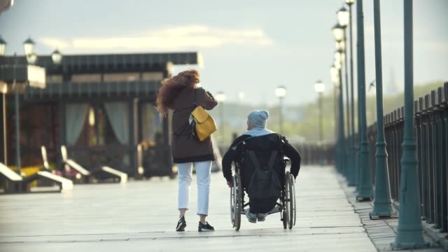 Young-happy-woman-with-disabled-man-in-a-wheelchair-walking-together-on-the-quay