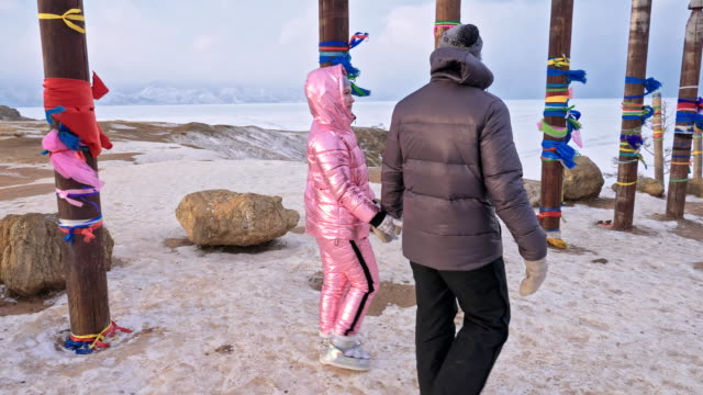 Joven-pareja-caminando-disfrutando-al-aire-libre-en-las-montañas-superiores-de-invierno.-Polos-de-Ritual-budista-con-cintas-de-colores-en-el-invierno-en-el-sitio-sagrado-del-lago-Baikal.