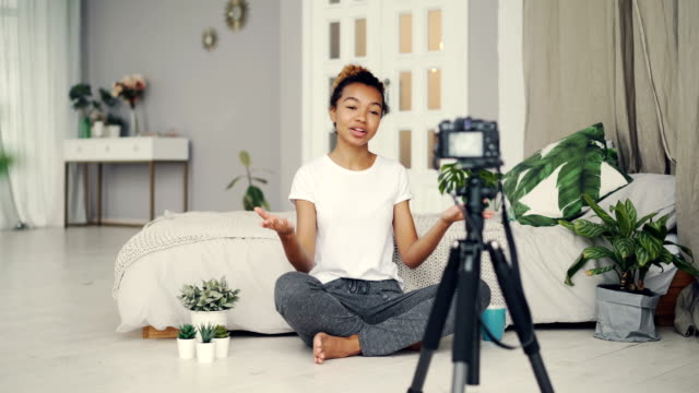 Gut-aussehende-afroamerikanische-Mädchen-kreativ-Blogger-zeichnet-Video-über-Pflanzen-im-Stock-ihrer-Wohnung-sitzen-und-reden,-Blick-in-die-Kamera-am-Stativ.