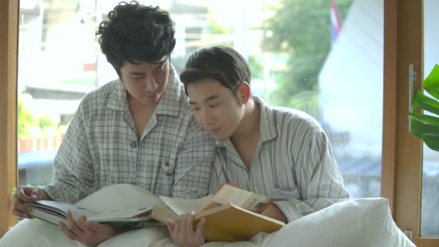 Asiático-gay-pareja-joven-sentada-en-el-alféizar-de-la-ventana-y-leyendo-el-libro-en-casa-en-la-mañana.