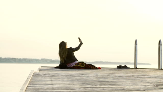 Ein-schönes-Mädchen-sitzen-auf-einem-Steg-am-Meer-unter-ein-Instagram-Storry-Selfie,-halten-Sie-das-Friedenszeichen-im-Sonnenuntergang