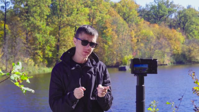 Brunette-man-blogger-in-sunglasses-on-river-bank-is-recording-video-for-vlog-using-camera