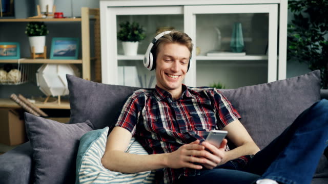El-joven-feliz-en-los-auriculares-está-escuchando-música-y-usando-el-teléfono-inteligente-en-casa