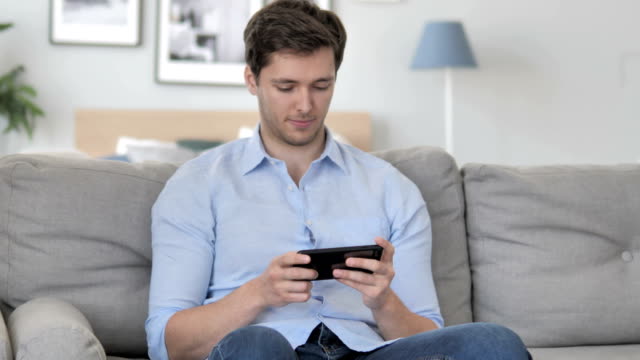 Hübsche-Young-Man-Cheering-Erfolg-auf-Smartphone-beim-Sitzen-auf-Couch