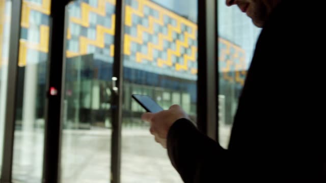Tracking-left-shot-of-unrecognizable-businessman-in-suit-texting-on-smartphone-while-walking-along-entrance-hall-in-office-building