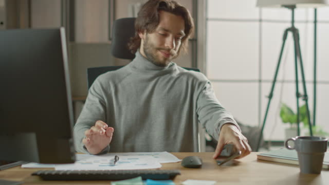 Handsome-Long-Haired-Entrepreneur-Sitzt-an-seinem-Schreibtisch-im-Büro-arbeitet-auf-Desktop-Computer,-Arbeiten-mit-Dokumenten,-Graphen.-Verwendet-Smartphone,-Social-Media-App,-Schreiben-von-E-Mails,-Messaging