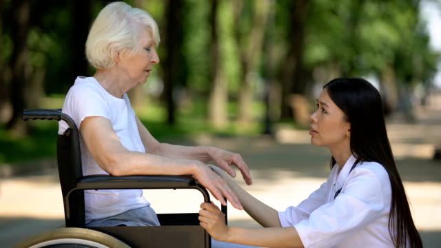 Freiwillige-Helferin-unterstützt-traurige-ältere-Frau-im-Rollstuhl-im-Krankenhauspark