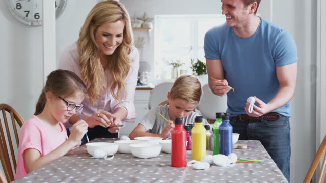 Familia-sentada-alrededor-de-la-mesa-de-la-cocina-con-los-padres-ayudando-a-los-niños-a-pintar-huevos-para-Semana-Santa---tiro-en-cámara-lenta