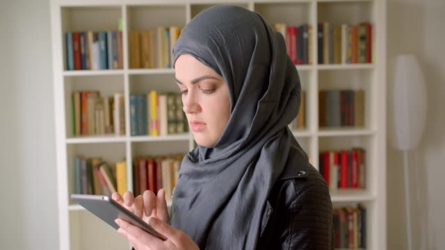Nahaufnahme-Porträt-von-jungen-attraktiven-muslimischen-Studentin-in-Hijab-mit-dem-Tablet-lächelnd-Blick-auf-die-Kamera-in-der-College-Bibliothek-in-Innenräumen