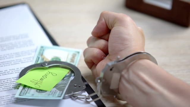 Male-hand-in-cuffs-and-dollars-with-alimony-sign-on-table,-obligatory-payment