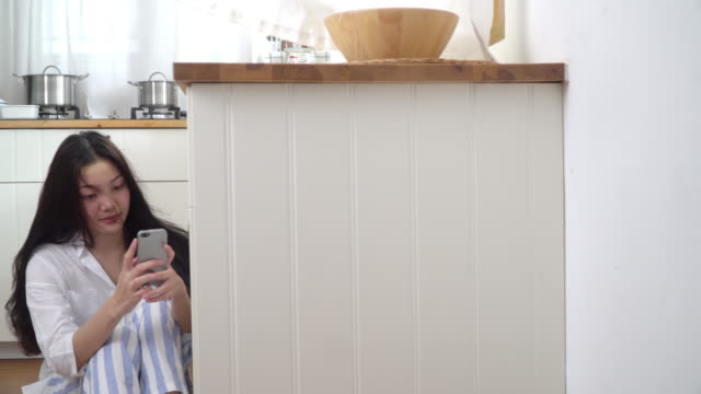 4K-Medium-Dolly-Schuss-von-jungen-asiatischen-Frau-sitzt-auf-Küchenboden-zu-Hause-mit-Smartphone-mit-drahtloser-Technologie-für-Social-Media-Kommunikation.-Asiatische-Teenager-Mädchen-genießen-auf-Küchenboden-mit-Smartphone.