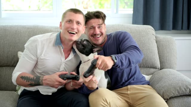 Gay-couple-relaxing-on-couch-with-dog-playing-games.-Dog-licking-face.