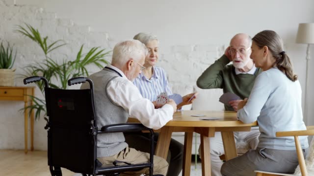 Grupo-de-cuatro-personas-mayores-jubiladas,-dos-hombres-y-dos-mujeres,-divirtiéndose-sentados-a-la-mesa-y-jugando-a-las-cartas-juntos-en-la-sala-común-de-residencia-de-ancianos,-rastreando-la-toma