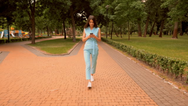 slim-businesswoman-using-smartphone-typing-message-in-blue-elegant-suit-walks-on-pedestrian-zone