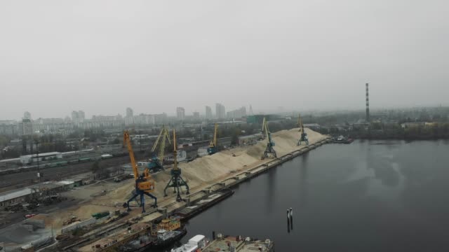 Luftaufnahme-der-Industriestadt-in-Smog-und-Nebel-mit-Baukränen,-die-an-Docks-arbeiten.-Sandkahn-und-Lastwagen-transportieren-Sand.