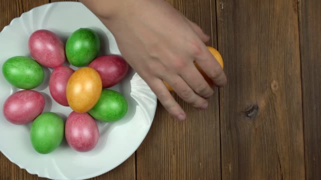 Una-persona-pone-huevos-de-pollo-multicolores-de-colores-en-un-plato,-fondo-de-madera,-vacaciones-de-Pascua,-espacio-de-copia,-casero