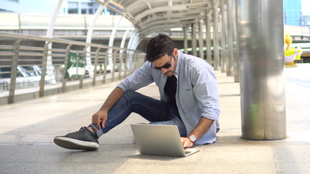 young-businessman-in-smart-casual-wear-sitting-on-street-using-laptop-and-typing-a-message-in-urban-city-outdoors.-freelancer-working-on-floor-outside-Chatting-to-customer.-blue-jeans