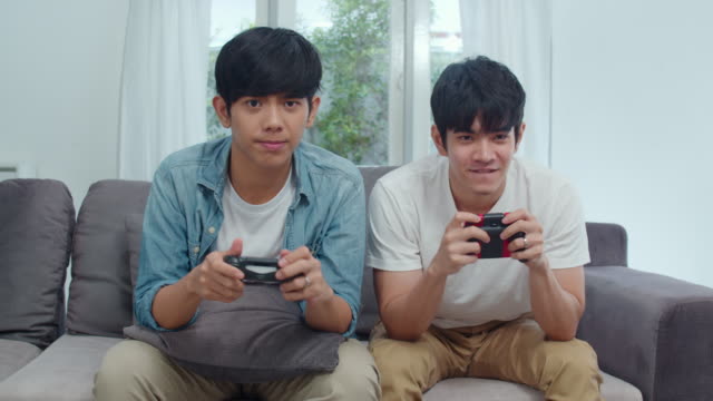 Joven-pareja-gay-asiática-jugar-juegos-en-casa,-adolescentes-coreanos-HOMBREs-LGBTQ-usando-joystick-que-tienen-divertido-momento-feliz-juntos-en-el-sofá-en-la-sala-de-estar-en-casa.