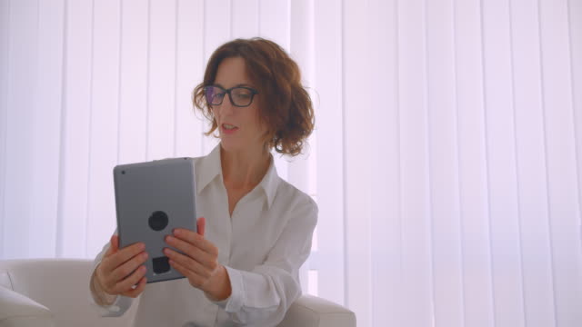 Nahaufnahme-Porträt-der-erwachsenen-Rotschopf-kaukasische-Geschäftsfrau-in-Brille-mit-einem-Videoanruf-auf-dem-Tablet-sitzen-dim-Sessel-im-weißen-Büro