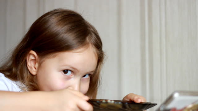 Kind-isst-Suppe-mit-einem-Löffel-und-schaut-Video-auf-einem-Smartphone.-Closeup-Baby-Mädchen-sitzt-am-Tisch-der-Hausmannskost-und-Essen-essen