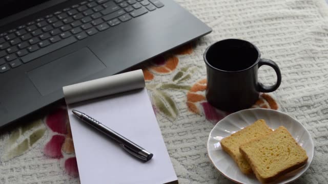 Breakfast-coffee-in-morning-sunlight-with-laptop-computer-black-color-pen-and-white-ruled-paper-notebook,-ceramic-cup-saucer-and-biscuit-on-top-office-place-working-desk-background.-Lifestyle-image.