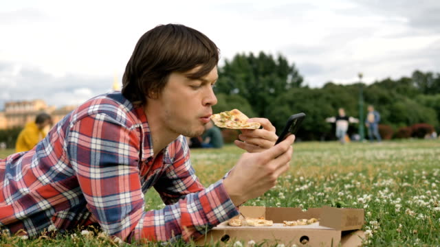 Mann-liegt-auf-dem-Gras-essen-Pizza-und-mit-einem-Smartphone-in-einem-Stadtpark-auf-die-Natur
