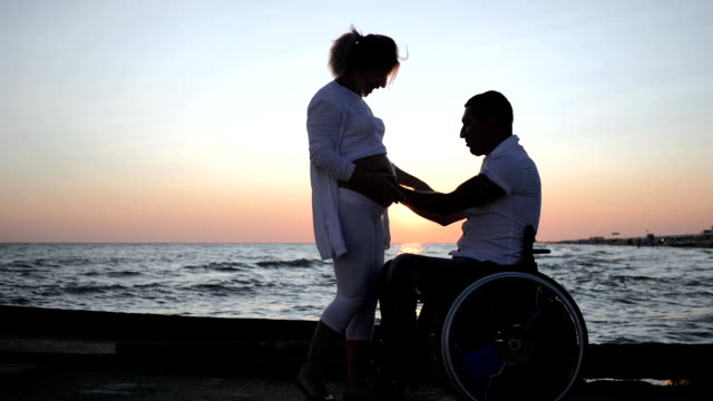 happy-disabled-man-caresses-woman-with-big-belly,-disabled-man-in-a-wheelchair-listens-to-the-belly-of-his-pregnant-wife-near-sea