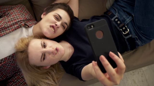 Two-young-lesbian-girls-lie-on-the-couch,-do-selfie-on-a-smartphone,-lovers,-lgbt,-young-couple,-teenagers-top-shot-60-fps