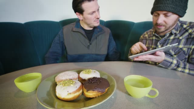 Zwei-Männer,-die-eine-wichtige-Profigeschäft-zufälligen-Begegnung-beim-Frühstück-Diner-Diskussion-Viewing-Tablet-Computer-Donuts-auf-Tisch