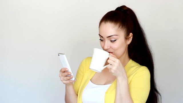 Portrait-of-a-beautiful-brunette-girl-who-drinks-a-hot-drink-from-a-cup-and-holds-a-mobile-phone-in-his-hand.-Young-woman-reads-social-network-and-looks-internet-on-smartphone-screen-and-smiles