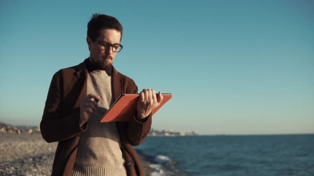 Hombre-con-barba-está-utilizando-ordenador-plano-sentado-en-la-orilla-del-mar-con-vista-panorámica-de-la-ciudad