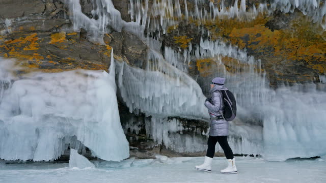 Travel-of-woman-on-ice-of-Lake-Baikal.-Trip-to-winter-island.-Girl-is-walking-at-foot-of-ice-rocks.-Traveler-looks-at-beautiful-ice-grotto.-Extreme-trek-and-walk.-Backpacker-is-resting-on-nature.